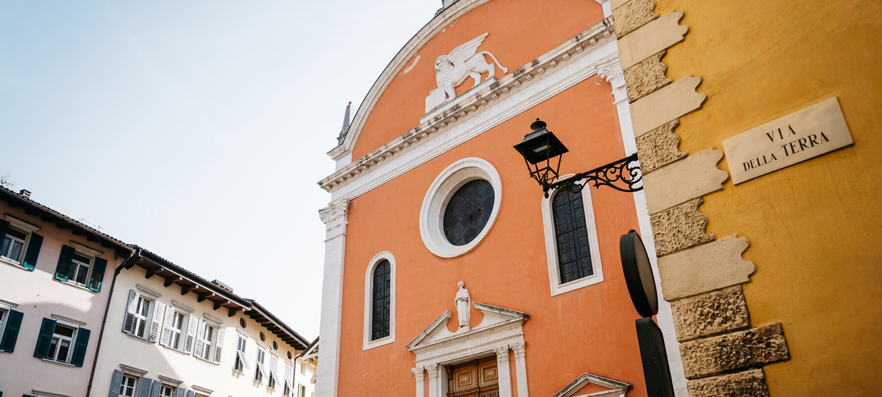 Façade of the Church of San Marco, in the historic center of Rovereto. The protagonists of the image are the colors: the orange of the church, the yellow ochre of the building in the foreground, on which “Via della Terra” is written, and the white and soft pink of the buildings in the background. Although not inhabited by people, it is a lively, joyful image.