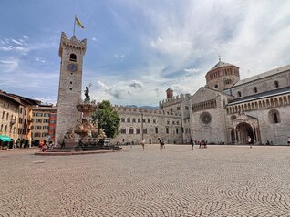 Piazza Duomo a Trento. Sullo sfondo, la Torre Civica, Palazzo Pretorio e la cattedrale di San Vigilio, Duomo della città. Al centro, la fontana del Nettuno. Dietro alla fontana svetta l’unico albero della piazza: un tiglio. La piazza, nell’immagine, è vissuta. Ci sono alcune persone sedute sui gradini della fontana, qualcuno sta scattando delle foto e un gruppetto è in posa davanti alla cattedrale per farsi fotografare. Tutti indossano abiti estivi. Il cielo azzurro è striato dal bianco di nuvole leggere.