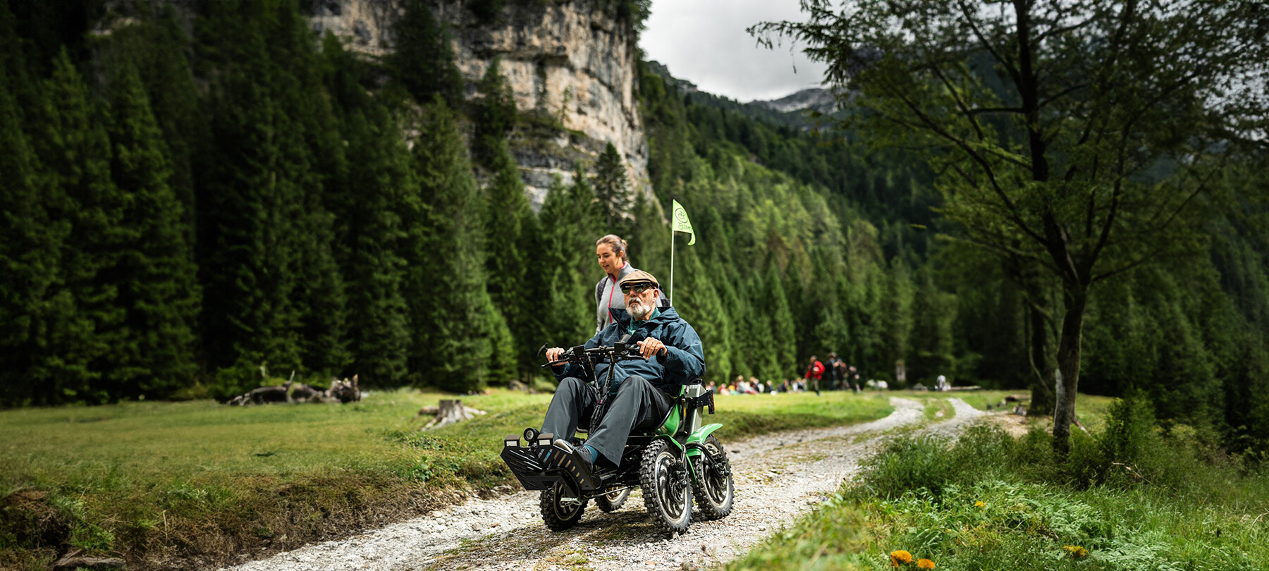 Ein Mann auf einem Quad bike entfernt sich vom Ort des Konzertes, das wahrscheinlich gerade zu Ende gegangen ist. Ein Mädchen geht neben ihm her und redet mit ihm. Der Himmel ist grau und die Luft muss frisch sein. Das Grün der Bäume im Hintergrund ist dunkel und intensiv.