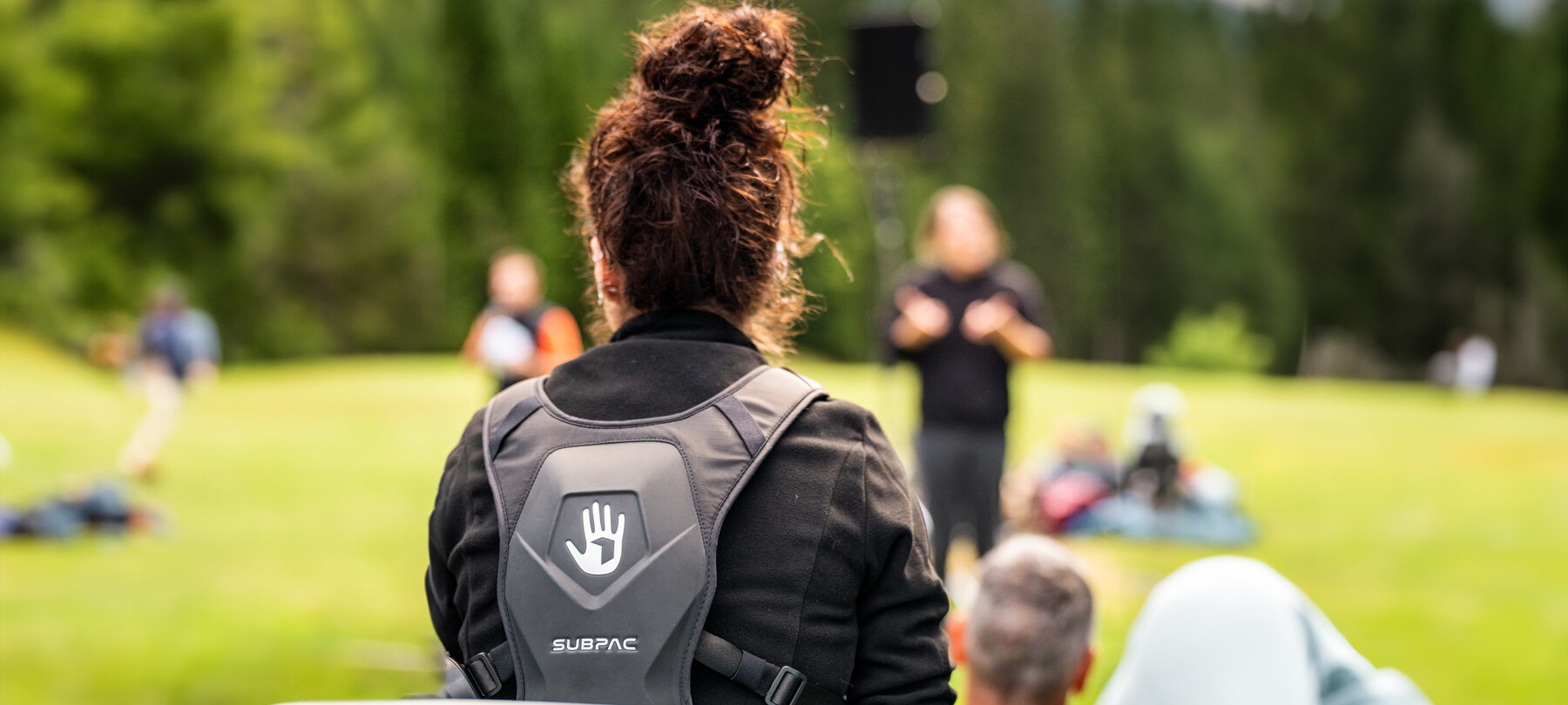Im Vordergrund trägt ein Mädchen, das man von hinten sieht, einen subpac-Rucksack. Vor ihr, im Bild verschwommen, treten Künstler auf, die Klänge erzeugen, deren Vibrationen sie spüren wird.