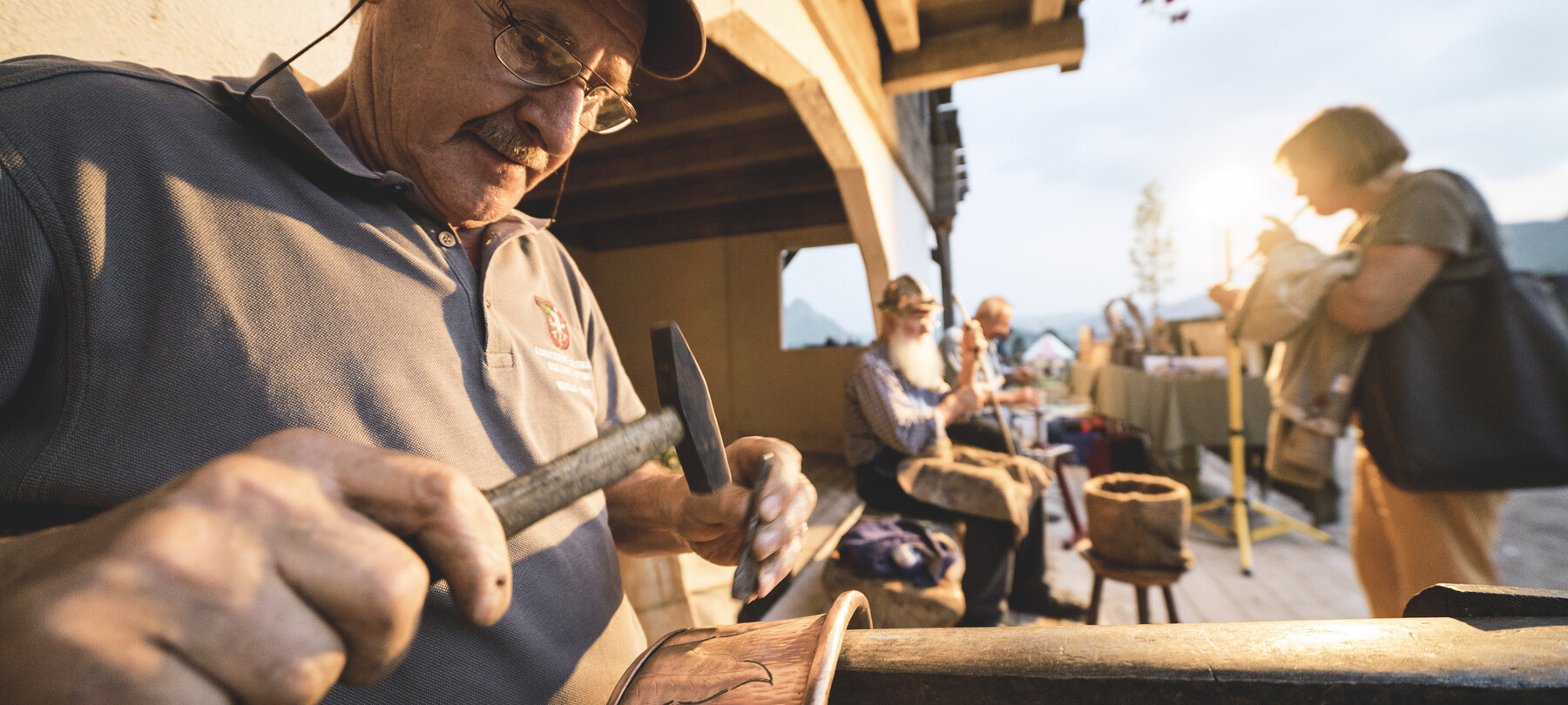 Artisans at work during the Tutti #Fuori event, a travelling festival of the Trentino Pro Loco associations. In the foreground, a man engraves decorations on a copper pot. The warm sunset light casts golden reflections on things and people.