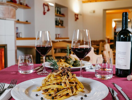 A table set in front of the fireplace at Malga Terlaga. In the foreground a plate of tagliatelle with mushrooms and two glasses of Teroldego Rotaliano, a red wine. A second dish can be glimpsed in the background.