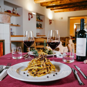 A table set in front of the fireplace at Malga Terlaga. In the foreground a plate of tagliatelle with mushrooms and two glasses of Teroldego Rotaliano, a red wine. A second dish can be glimpsed in the background.