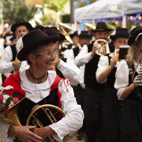 The photograph portrays a group of musicians in traditional costume, likely during a folk festival. In the foreground, an elderly woman smiles, wearing a red and white dress with a black hat, holding a golden horn and a bouquet of red and white flowers. In the background, other musicians in similar attire play instruments and wear dark hats. The atmosphere is festive and cheerful, with lights and decorations suggesting an outdoor event in a square or street. The image conveys a strong sense of tradition, community, and shared joy.