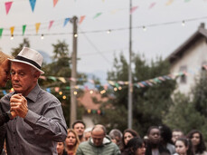 A couple dances tenderly in the town square during the festival, surrounded by onlookers of all ages. String lights and colorful flags decorate the scene. The moment conveys a sense of community, warmth, and togetherness.