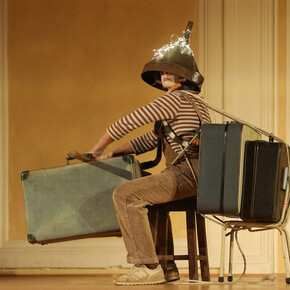 L'immagine mostra la protagonista durante lo spettacolo mentre è in partenza con le sue valigie, un imbuto in testa e delle lucine sopra