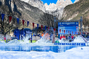 Athleten treten im Rückenschwimmen in einem Freibad während der Eisschwimm-Weltmeisterschaften gegeneinander an. Die Aufnahme auf Wasserhöhe zeigt das von Eis umgebene Becken und die imposanten Dolomitenberge als Kulisse für den Wettbewerb.