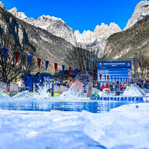 Athletes compete in the backstroke in an outdoor pool during the Ice Swimming World Championships. The low-angle shot, taken at water level, shows the pool surrounded by ice and the imposing Dolomite mountains providing a backdrop for the competition.