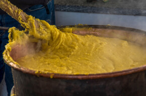 La foto cattura il primo piano di un paiolo colmo di polenta mentre un uomo mescola con un grande bastone in legno