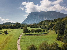 Veduta aerea di un sentiero sterrato che serpeggia tra ampi prati verdi e boschetti a Fai della Paganella. Lungo il percorso si vedono alcuni gruppi di persone che camminano, mentre sullo sfondo si staglia la Paganella.