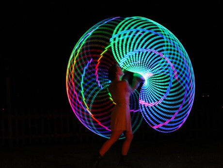 Nell’immagine si vede una persona che si esibisce di notte all’aperto, creando con il movimento grandi cerchi colorati e luminosi che formano un effetto a spirale nel buio. I colori accesi — blu, verde, rosso e giallo — illuminano la scena come un disegno di luce in movimento.