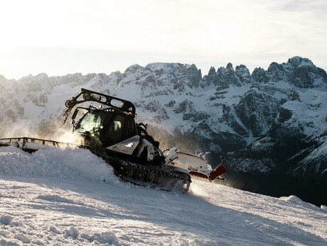  A snowcat is at work on a snowy slope, lifting fresh snow to the sides while levelling the ground. In the background are the snow-capped Brenta Dolomites, illuminated by the diffuse light of the sky, which appears clear with light clouds.