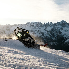  A snowcat is at work on a snowy slope, lifting fresh snow to the sides while levelling the ground. In the background are the snow-capped Brenta Dolomites, illuminated by the diffuse light of the sky, which appears clear with light clouds.