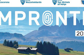 Paesaggio alpino con prati verdi, baite in legno e boschi ai piedi di una catena montuosa, immagine simbolo della rassegna culturale Impronte 2026