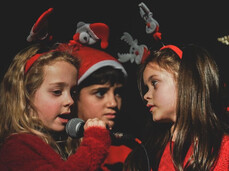 A close-up, half-body portrait shows three children singing together on a dark stage. The two children on the sides are dressed in red outfits and are wearing Christmas headbands, one with reindeer and the other with a miniature Santa Claus. The child in the center is wearing a Santa hat. The two girls on the sides are holding microphones and looking at each other while singing