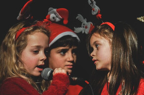 A close-up, half-body portrait shows three children singing together on a dark stage. The two children on the sides are dressed in red outfits and are wearing Christmas headbands, one with reindeer and the other with a miniature Santa Claus. The child in the center is wearing a Santa hat. The two girls on the sides are holding microphones and looking at each other while singing