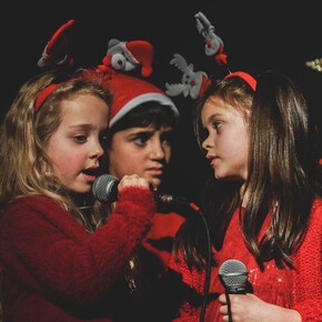 A close-up, half-body portrait shows three children singing together on a dark stage. The two children on the sides are dressed in red outfits and are wearing Christmas headbands, one with reindeer and the other with a miniature Santa Claus. The child in the center is wearing a Santa hat. The two girls on the sides are holding microphones and looking at each other while singing