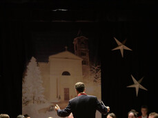 Un'orchestra è ritratta su un palco. Il direttore, di spalle, è al centro e dirige i musicisti seduti, la maggior parte dei quali indossa gilet rossi e camicie bianche. Gli strumenti includono clarinetti, sassofoni e una tuba. Sullo sfondo nero è appeso un telo raffigurante una chiesa bianca con un campanile e alberi innevati, affiancato da grandi decorazioni a forma di stella. L'atmosfera è quella di un concerto o di un'esibizione natalizia al chiuso.