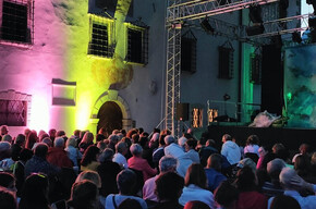 The image shows an audience of about 200 people seated, waiting for the show to begin. The chairs are arranged in the courtyard of a 19th-century building, illuminated by yellow and pink lights. On the stage, there are structures that are likely part of the upcoming performance. The people are smiling and eagerly anticipating the start of the show.