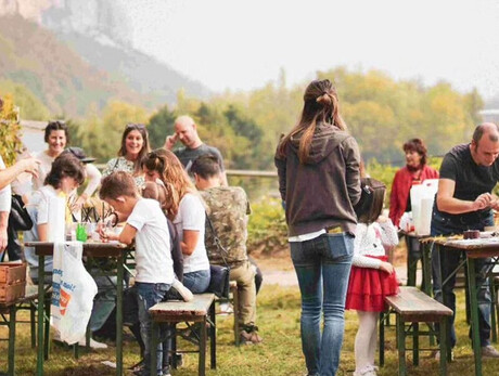 A group of children and adults take part in an outdoor activity surrounded by nature, with craft tables, games, and a warm, communal atmosphere. The event feels friendly and relaxed, ideal for a local gathering. Hills and vineyards can be seen in the background.