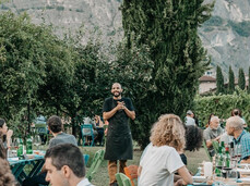 A large group of people is seated outdoors among the vineyards, while a man in the center, likely the host, welcomes guests with a smile. The atmosphere is warm and convivial, surrounded by nature and mountains.