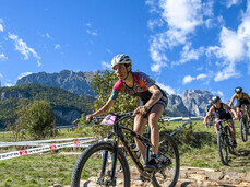 Three participants from X-Terra ride their mountain bikes over wooden logs on the beach of Lake Molveno. In the background you can see the Brenta Dolomites
