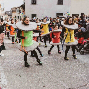 This photo captures a Carnival parade in Roveré della Luna, featuring participants dressed as colorful spools of thread. The people, including adults and children, wear creative costumes with details like threads and sewing pins. The crowd applauds and takes photos, creating a festive atmosphere. In the background, traditional village houses and other masked participants can be seen. The ground is covered in confetti, adding vibrancy to the event. The image perfectly captures the joy and energy of the celebration.
