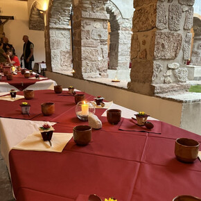 The photo shows a long burgundy table carefully set up under the stone porch of the ancient cloister of the Franciscan convent. The atmosphere is intimate and serene, with lit candles, clay glasses, and floral decorations. In the background, people can be seen conversing near a statue of the crucified Christ.