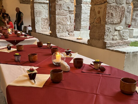 The photo shows a long burgundy table carefully set up under the stone porch of the ancient cloister of the Franciscan convent. The atmosphere is intimate and serene, with lit candles, clay glasses, and floral decorations. In the background, people can be seen conversing near a statue of the crucified Christ.
