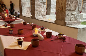 The photo shows a long burgundy table carefully set up under the stone porch of the ancient cloister of the Franciscan convent. The atmosphere is intimate and serene, with lit candles, clay glasses, and floral decorations. In the background, people can be seen conversing near a statue of the crucified Christ.