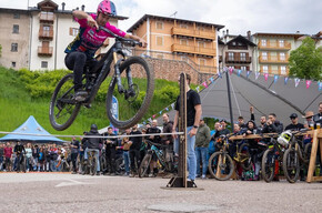 A young biker with a pink helmet jumps nimbly over a pole during a competition. She wears a fuchsia T-shirt and black trousers. Around her, a large group of spectators watches the action with enthusiasm. The background shows mountain-style buildings, a green hill and some decorations creating a lively atmosphere.