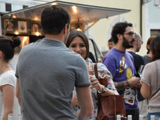 Persone che chiacchierano e degustano vino in piazza durante un evento estivo, tavoli con bottiglie e bicchieri.