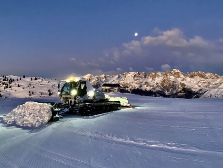 A snowcat is standing on a well groomed slope, its headlights illuminating the snow in front of it. In the background we see the snow-capped Brenta Dolomites under a twilight sky, with the moon visible among the scattered clouds.