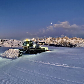 A snowcat is standing on a well groomed slope, its headlights illuminating the snow in front of it. In the background we see the snow-capped Brenta Dolomites under a twilight sky, with the moon visible among the scattered clouds.