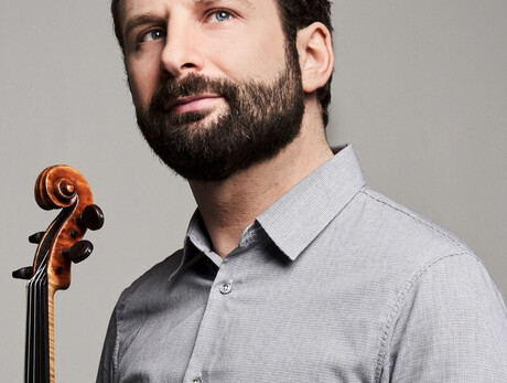 A man with a dark beard and micro-patterned gray shirt looks up towards the right against a gray background. At the bottom left, the neck and scroll of a violin are held near him.