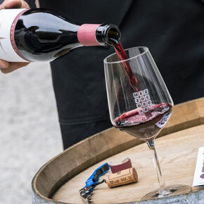 Das Bild zeigt im Vordergrund eine Flasche Teroldego Rotaliano, die in ein Glas eingeschenkt wird, das auf einem Holzfass steht. Höchstwahrscheinlich wird die Flasche von einem Sommelier auf der Veranstaltung eingeschenkt.