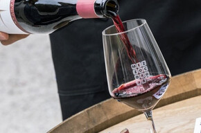 The image shows a bottle of Teroldego Rotaliano in the foreground being poured into a glass placed on a wooden barrel. Most likely, the bottle is being poured by a sommelier attending the event.