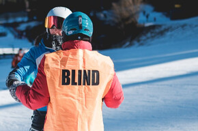 A visually impaired skier, identifiable by the orange bib with the word "BLIND" on it, is guided by a companion on a snowy ski slope.