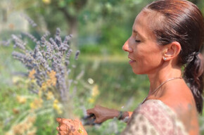 The photo portrays Annachiara Zecchetto, a nature guide, while she collects helichrysum and lavender plants for the Passo dopo Passo event that will be held in ACQUAin on July 26. Annachiara has an olive tree and a meadow with herbs in the background. She is holding a bunch of lavender and helichrysum. Her face conveys serenity. She is wearing a light and loose dress with a colorful print. The sun is high.