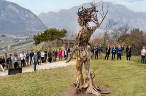Un gruppo di persone è riunito all’aperto su un prato verde, disposto in cerchio attorno a una grande scultura in legno raffigurante una figura umana stilizzata, realizzata con rami intrecciati e radici. Sullo sfondo si estende un ampio panorama di monti e vallate, con i vigneti e le case della Piana Rotaliana visibili in lontananza. Il cielo è parzialmente nuvoloso, e la luce morbida del giorno illumina la scena, creando un’atmosfera serena e contemplativa.