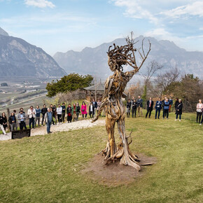 Un gruppo di persone è riunito all’aperto su un prato verde, disposto in cerchio attorno a una grande scultura in legno raffigurante una figura umana stilizzata, realizzata con rami intrecciati e radici. Sullo sfondo si estende un ampio panorama di monti e vallate, con i vigneti e le case della Piana Rotaliana visibili in lontananza. Il cielo è parzialmente nuvoloso, e la luce morbida del giorno illumina la scena, creando un’atmosfera serena e contemplativa.