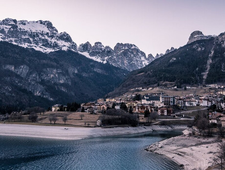 A calm, deep blue lake occupies the lower part of the picture, surrounded by a beach of light-coloured pebbles. In the background is Molveno: houses with dark roofs, low buildings and a visible church with a bell tower. Behind the village, the Brenta Dolomites rise imposingly, covered by dark forests and some snow-capped peaks. The sky is clear and fades to light tones. At sunset, the atmosphere is calm and cold, typical of a winter day.