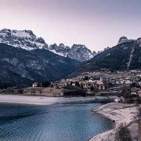 A calm, deep blue lake occupies the lower part of the picture, surrounded by a beach of light-coloured pebbles. In the background is Molveno: houses with dark roofs, low buildings and a visible church with a bell tower. Behind the village, the Brenta Dolomites rise imposingly, covered by dark forests and some snow-capped peaks. The sky is clear and fades to light tones. At sunset, the atmosphere is calm and cold, typical of a winter day.