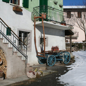 L'immagine mostra la facciata di una casa con una scala esterna in cemento, decorata con legna impilata sotto l’arco della scala. Accanto, un carro di legno con ruote azzurre è posizionato vicino al muro, decorato con vasi di piante. Sullo sfondo c'è un cortile innevato e un edificio con pareti rosa chiaro, circondato da alberi spogli.
