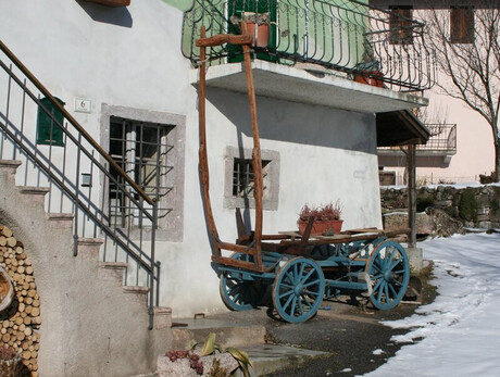 The picture shows the façade of a house with an exterior concrete staircase, decorated with wood stacked under the arch of the staircase. Next to it, a wooden wagon with blue wheels stands near the wall, decorated with pots of plants. In the background is a snowy courtyard and a building with light pink walls, surrounded by bare trees.