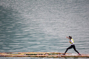 An athlete, during the XWarrior race in Molveno, is balancing while running across a narrow, floating walkway made of logs.