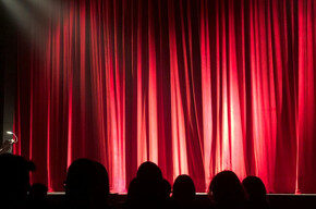 Palco teatrale con sipario rosso chiuso e luci di scena, pubblico seduto in platea.