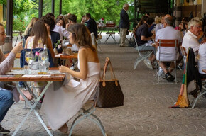 Many people seated at tables are enjoying a summer aperitif under the veranda of the Endrizzi Winery. The atmosphere is elegant yet welcoming. String lights on the roof create a warm ambiance. Around the veranda, a vineyard glows in vibrant green. Glasses of wine are served with delicious appetizers.