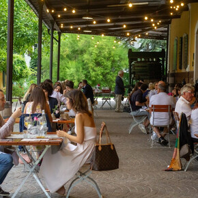 Many people seated at tables are enjoying a summer aperitif under the veranda of the Endrizzi Winery. The atmosphere is elegant yet welcoming. String lights on the roof create a warm ambiance. Around the veranda, a vineyard glows in vibrant green. Glasses of wine are served with delicious appetizers.
