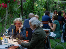 A romantic and elegant dinner among the vineyard rows, under soft lights and surrounded by greenery. Couples and groups of friends chat while enjoying wine and gourmet dishes. The atmosphere is relaxed, intimate, and immersed in nature.